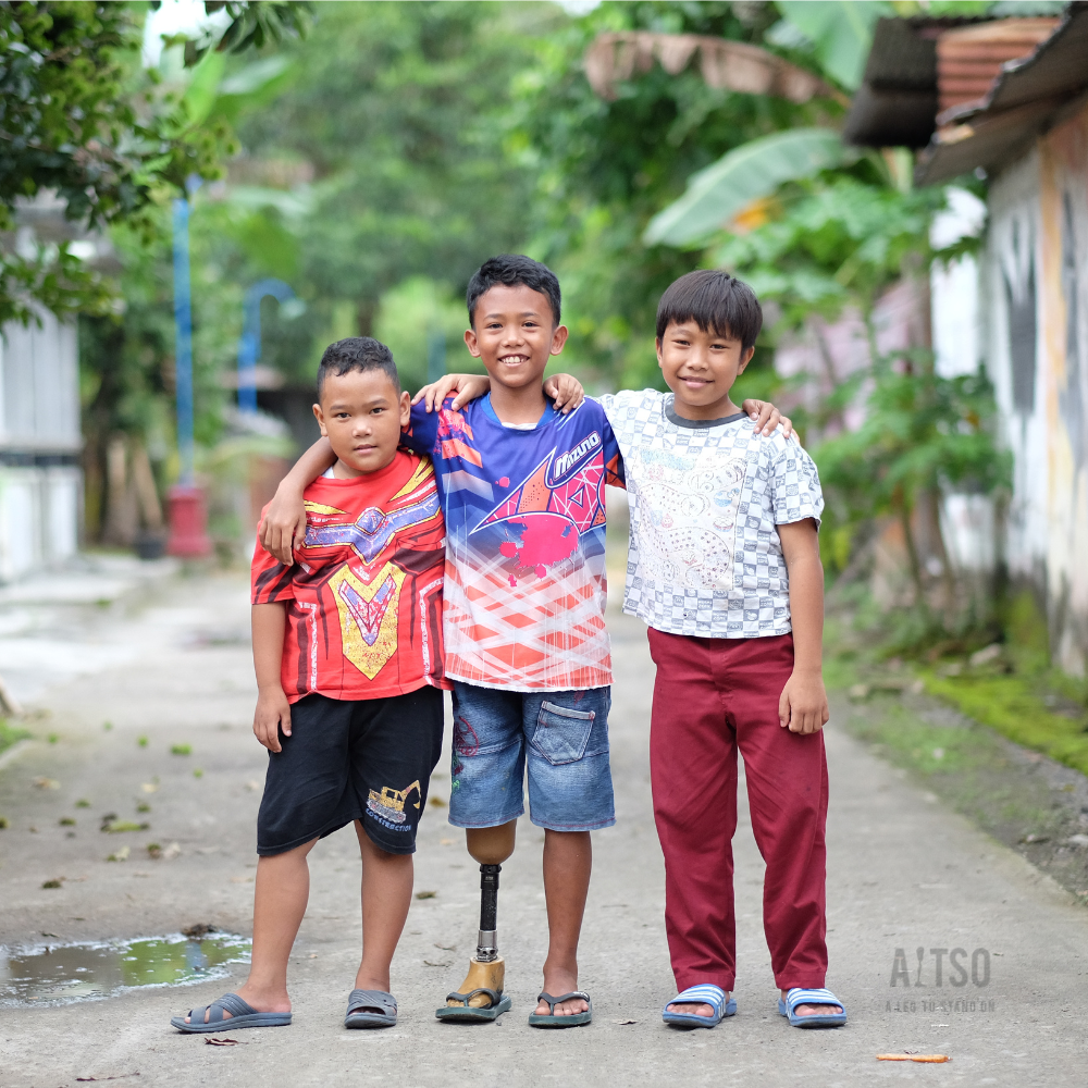 Three children stand outdoors with their arms around each other, smiling; one child uses a prosthetic leg