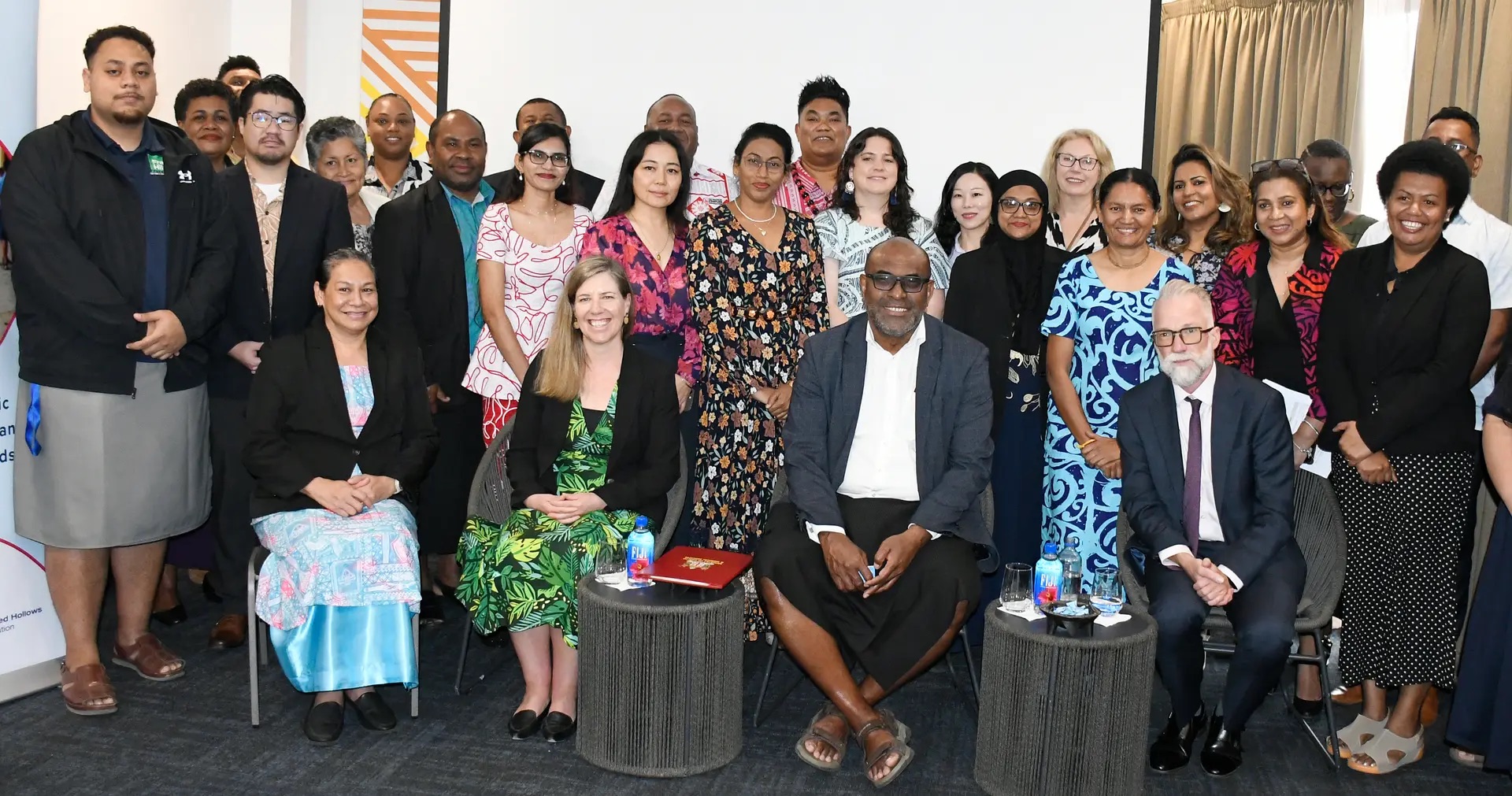 A group photo of diverse attendees and presenters posing together indoors at an event, with four key representatives seated in the front row and a large group standing behind them.