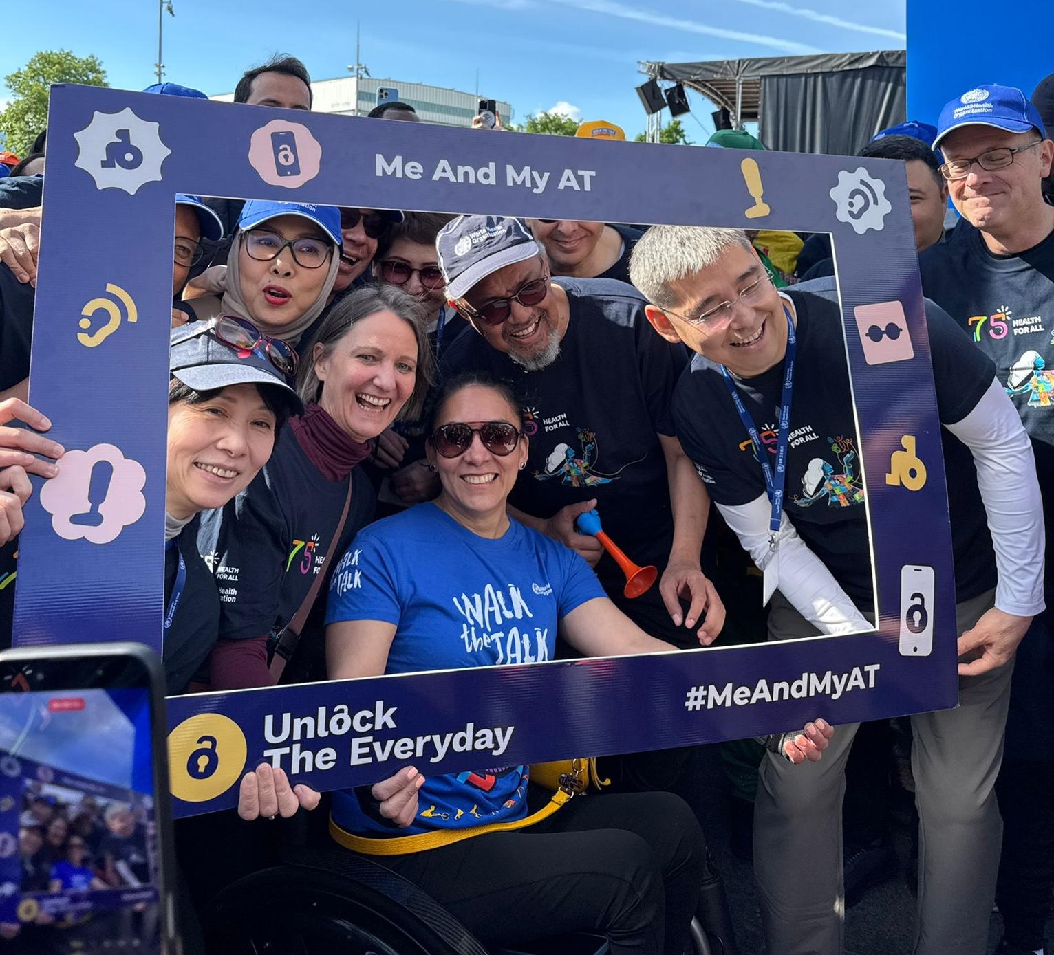 A group of smiling participants pose together behind a "#MeAndMyAT" and "Unlock The Everyday" campaign photo frame at a WHO Walk the Talk event