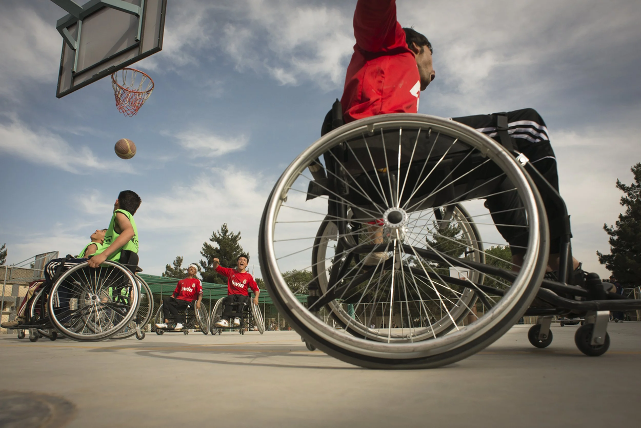 Wheelchair basketball players competing outdoors, one athlete shooting toward the hoop while teammates watch