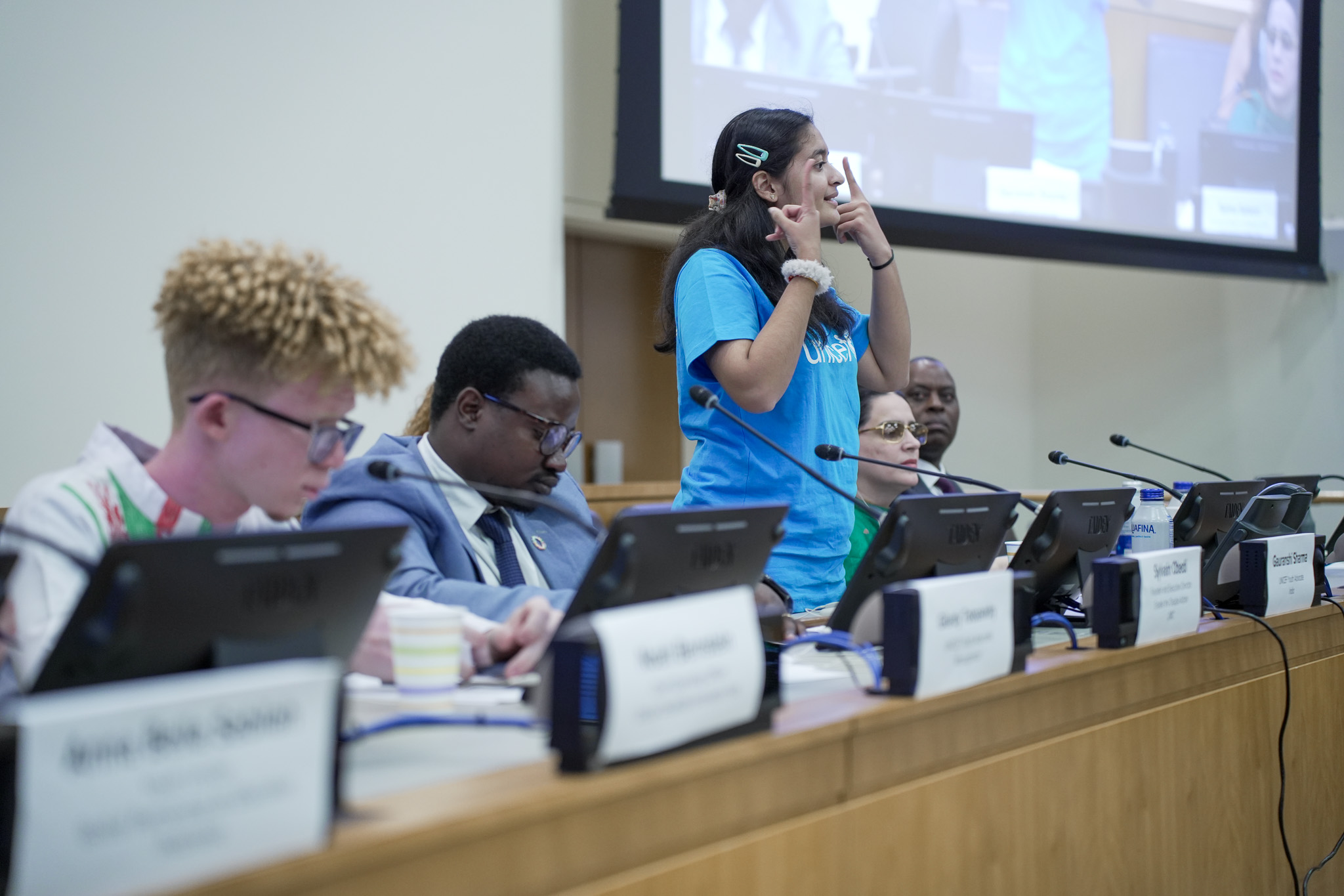 “A young woman uses sign language to address a panel during an international conference, with seated delegates and microphones in front of her