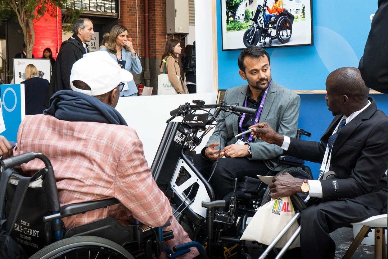 Three people in wheelchairs discuss and examine an assistive mobility device at an exhibition booth.”