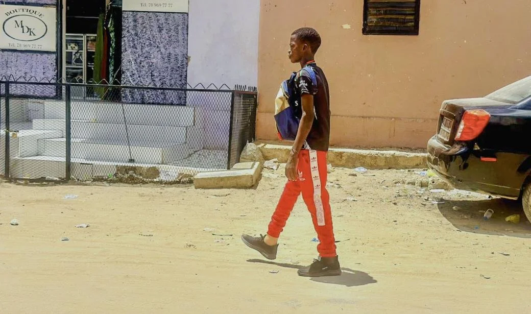 A young person walks along a dusty street in an urban neighbourhood, wearing a backpack and red trousers