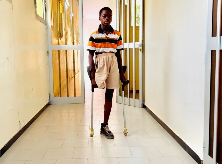A young boy walks down a corridor using forearm crutches, moving independently through a building hallway.