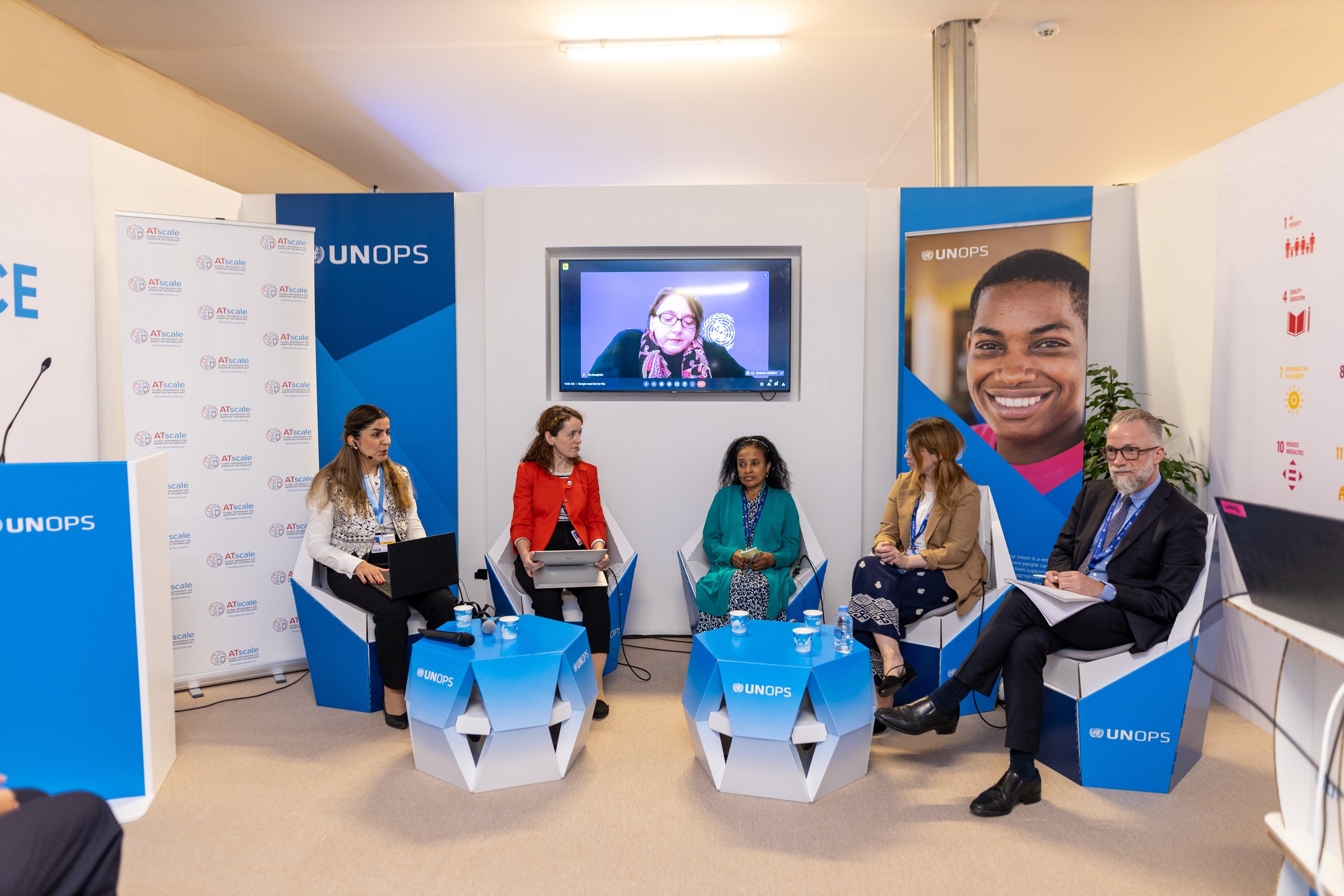 Panel discussion on assistive technology at a UNOPS event, with five speakers seated on stage and one participant joining remotely on screen.