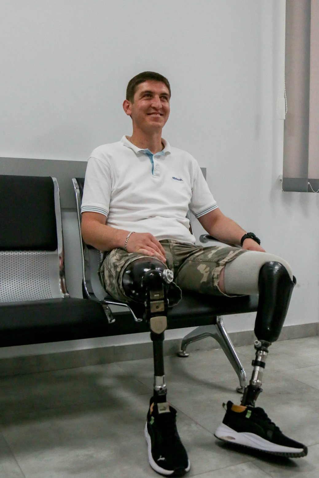 Man with bilateral prosthetic legs sits on a waiting-room bench, smiling, wearing a white polo shirt and shorts