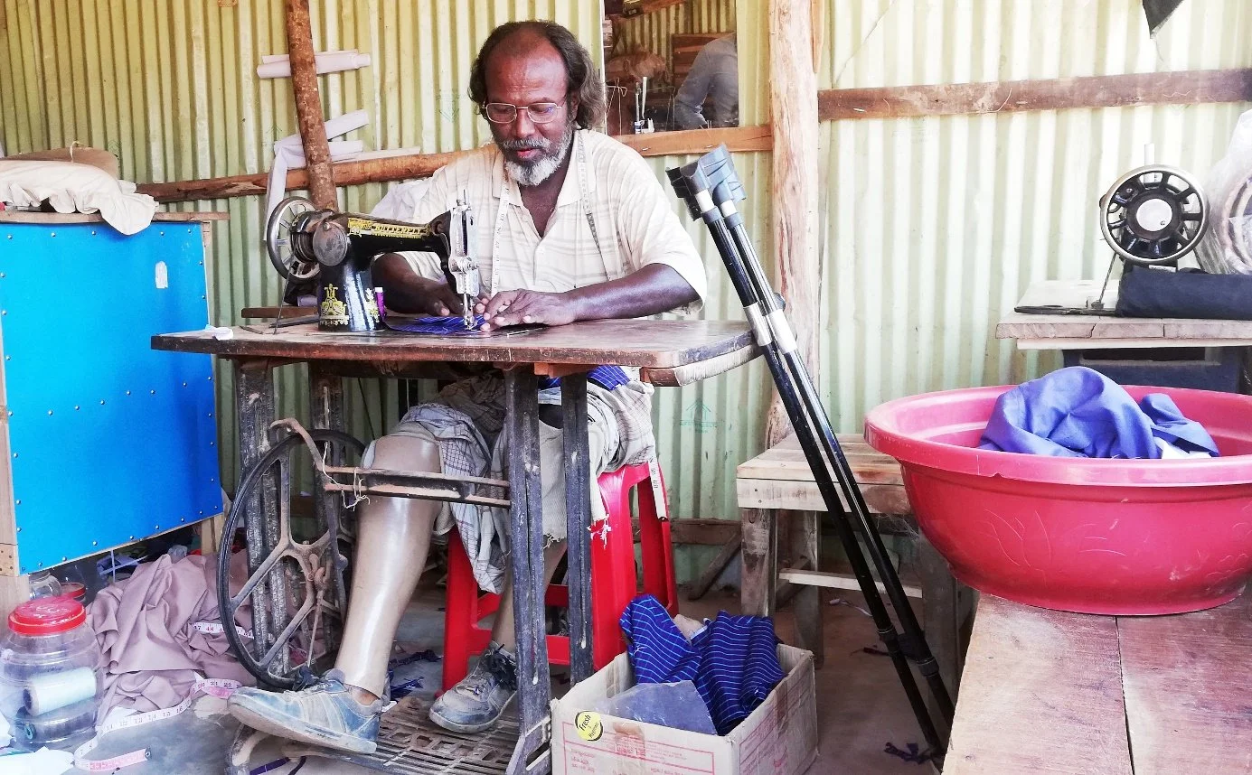 Man with a prosthetic leg uses a treadle sewing machine in a small workshop, with crutches resting nearby.