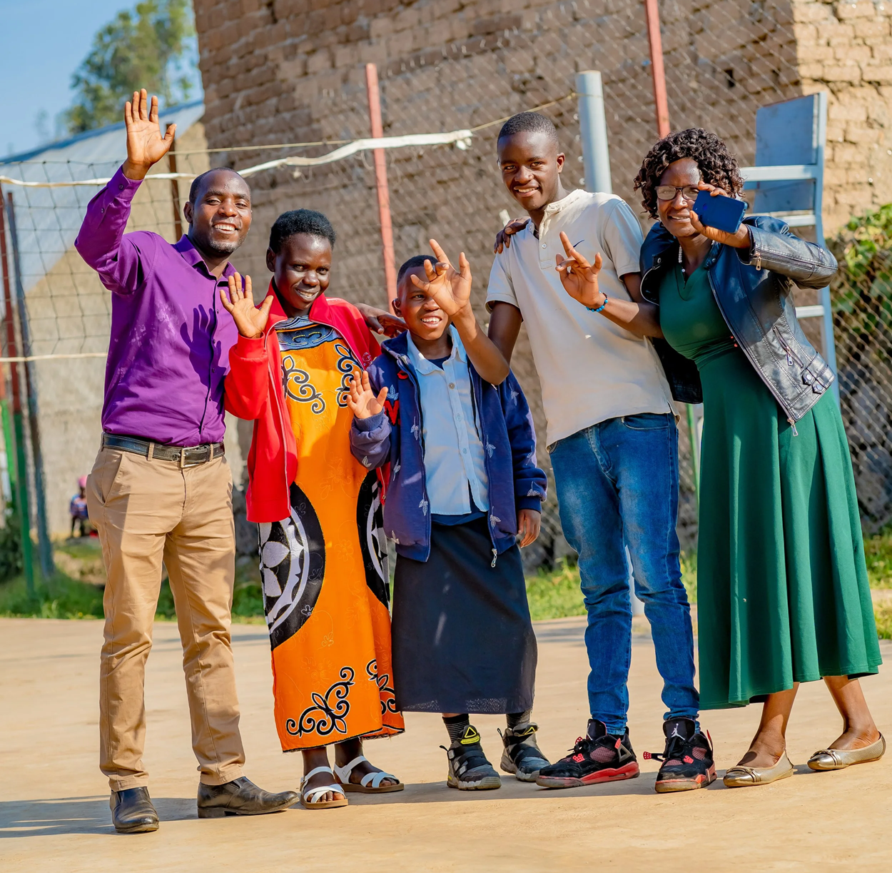 Six people of different ages stand outdoors in front of a brick building, smiling and waving at the camera in a community setting.