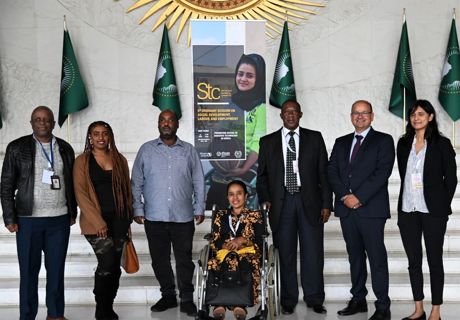 Group of delegates posing in a formal hall with African Union flags; one woman seated in a wheelchair at center, standing in front of a banner on access to assistive technology and social development.