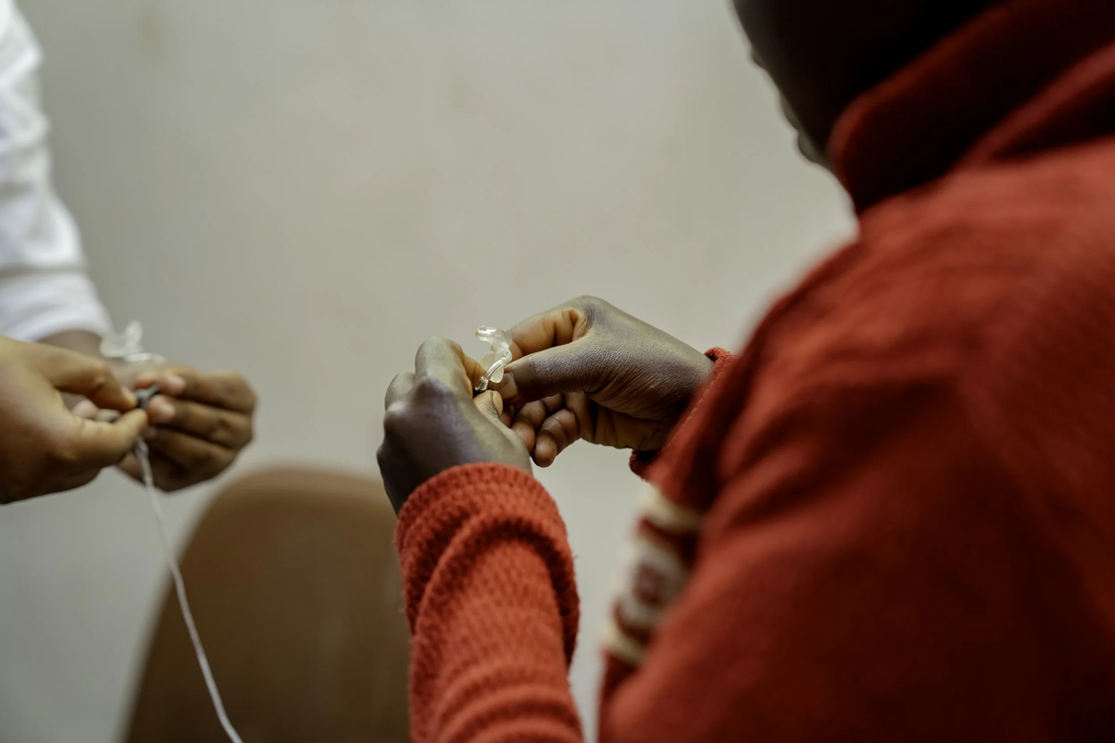 Close-up of hands fitting or adjusting a small hearing device, with assistance from another person.