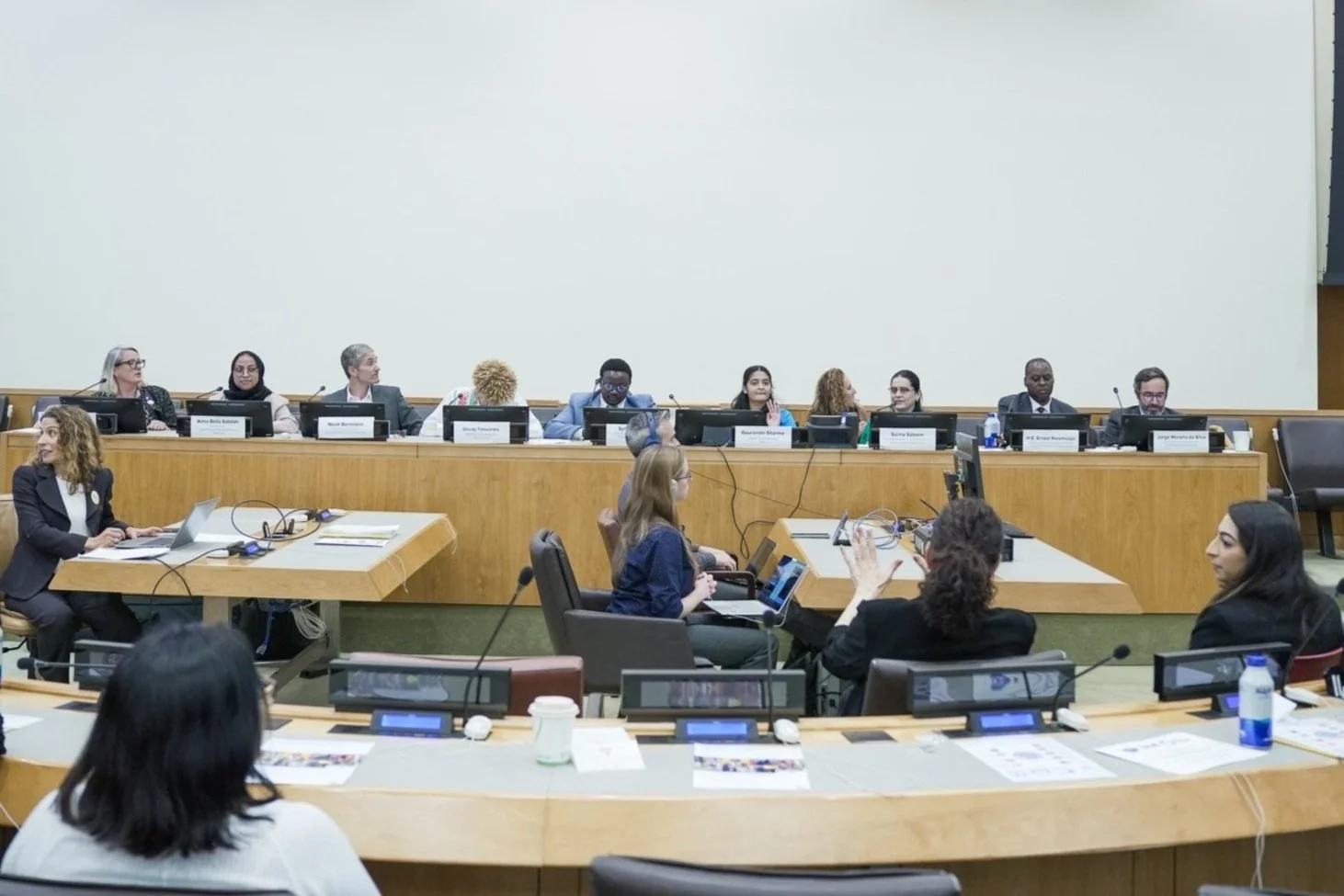 Wide view of a formal panel discussion in a large conference room, with multiple speakers seated behind desks and participants engaging in discussion.