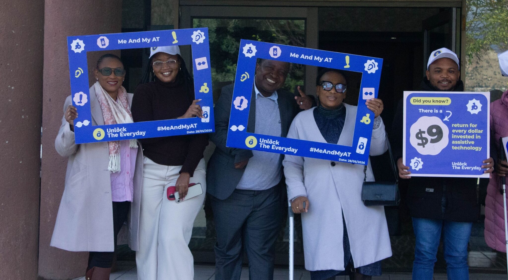 Advocates pose outdoors holding blue "#MeAndMyAT" photo frames and an "Unlock The Everyday" campaign sign highlighting investment in assistive technology.