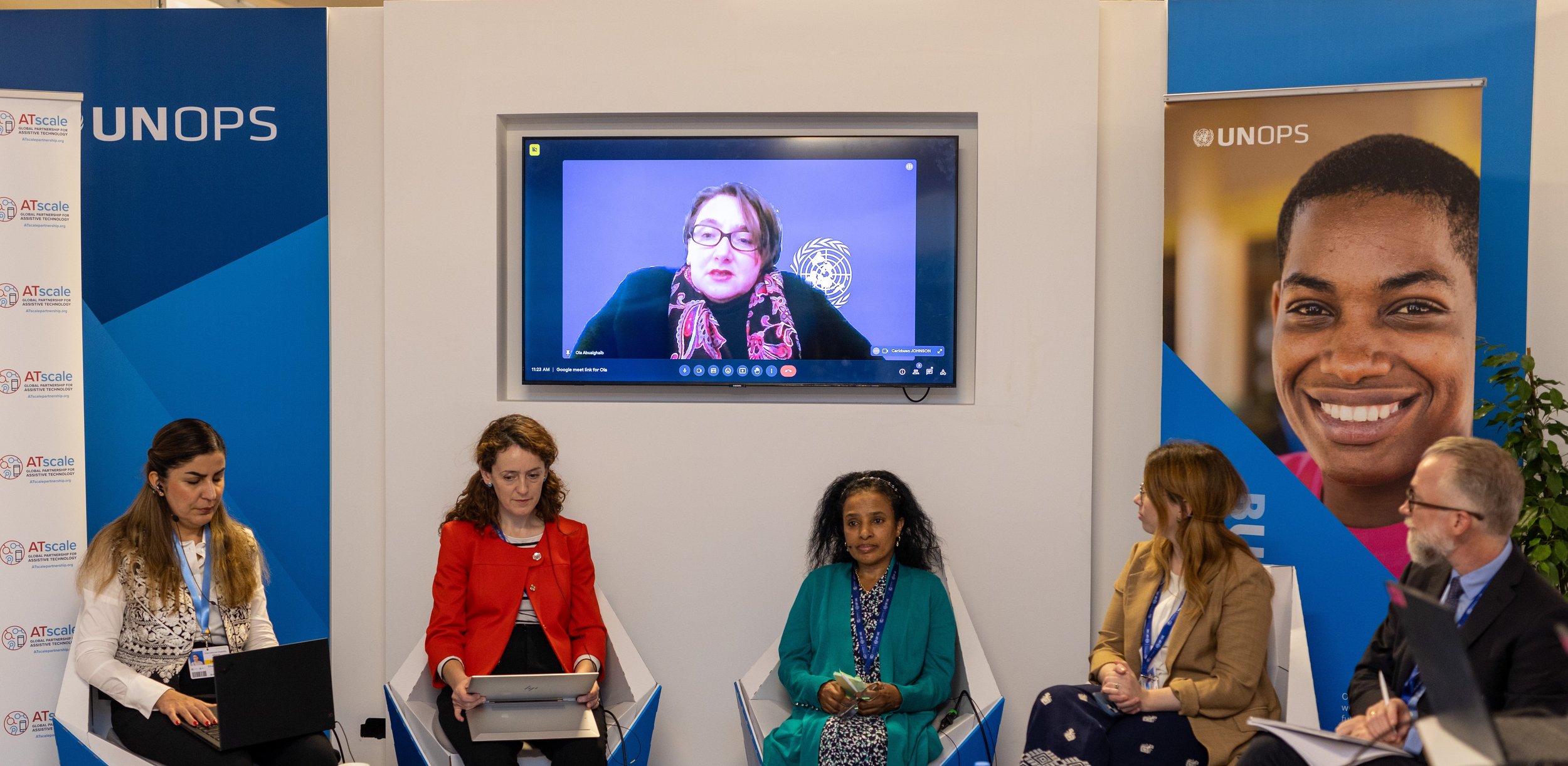 Panel discussion on assistive technology at a UNOPS event, with five speakers seated on stage and one participant joining remotely on screen.