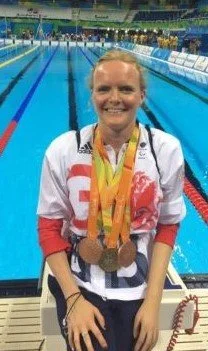 Susannah Rodgers MBE, Paralympic Gold and Bronze Medallist in Swimming, woman athlete wearing multiple medals sits beside a swimming pool, dressed in a Team Great Britain jacket