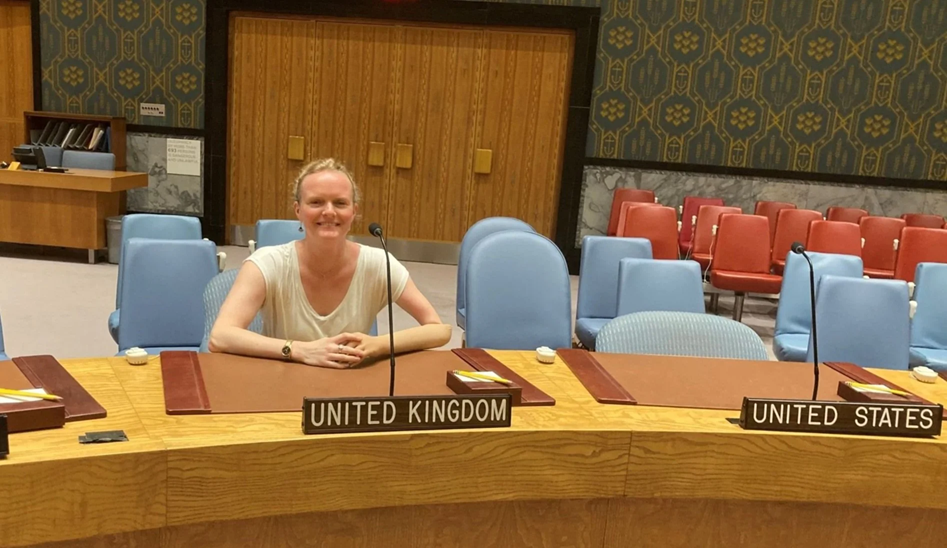 A woman seated at a United Nations conference desk labeled ‘United Kingdom,’ smiling in an empty meeting room with microphones and chairs visible.