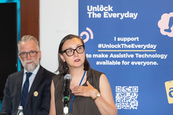 A woman speaks at a podium during an event, with an ‘Unlock the Everyday’ banner about assistive technology behind her and another speaker seated nearby.