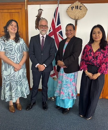 Four people standing side-by-side in formal attire, posing indoors in front of the Fijian flag and traditional woven wall decor.