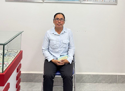 A woman wearing glasses sits on a chair inside a vision center, holding an eyeglass case, with anatomical eye charts displayed on the wall behind her.