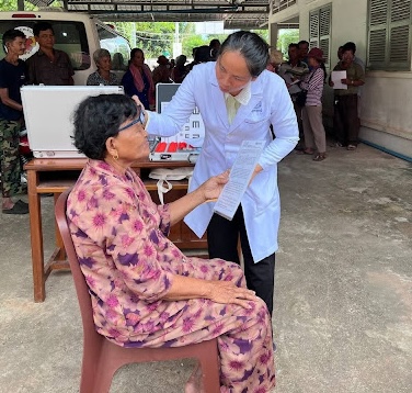 A healthcare worker in a white lab coat conducts an eye examination on a seated woman at an outdoor mobile health clinic.