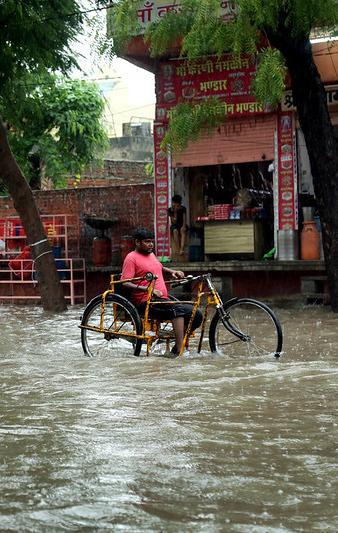 Man using a hand-powered tricycle moves through floodwater on a city street.