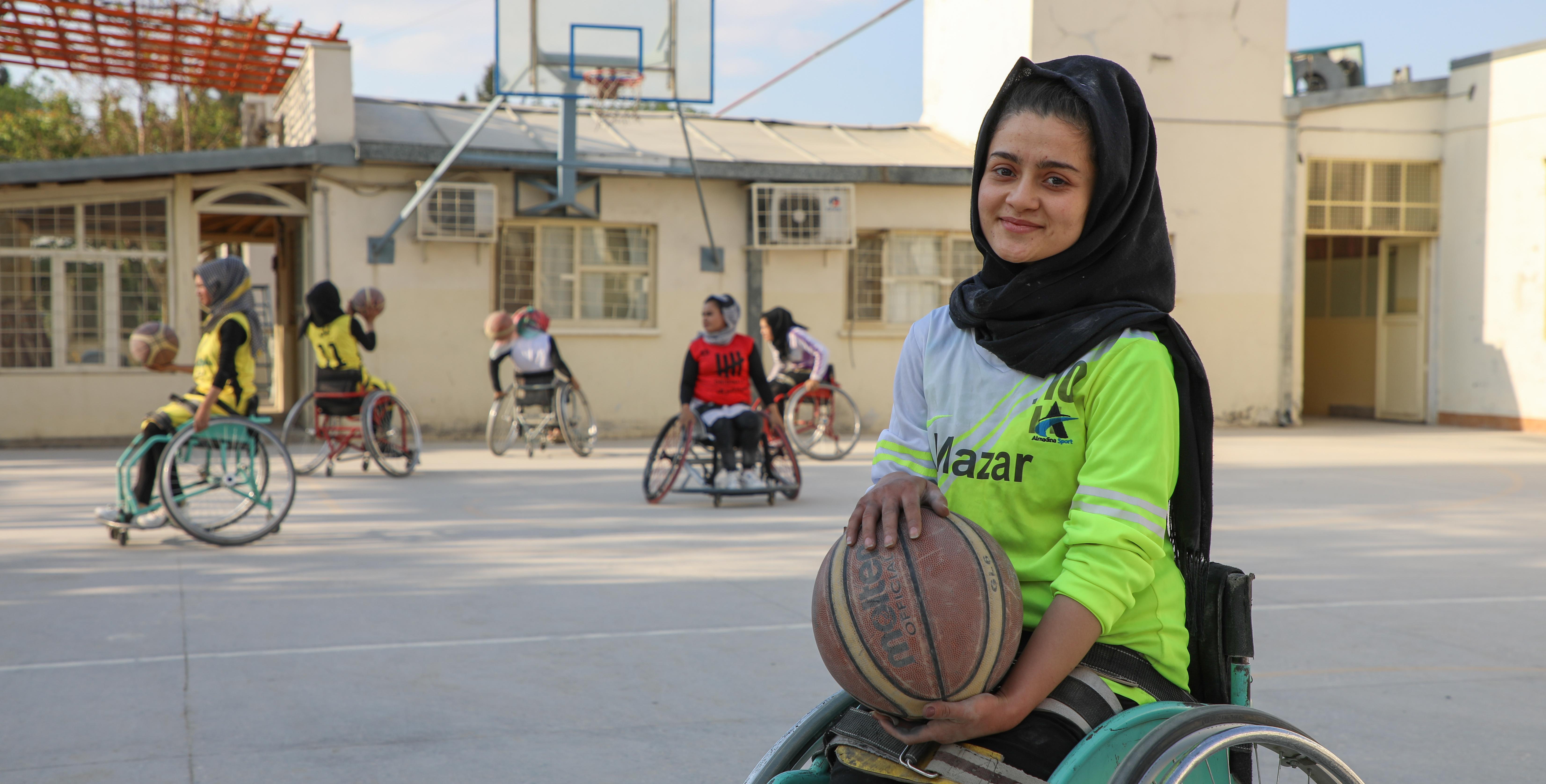 A young woman using a wheelchair holds a basketball on an outdoor court, with others playing wheelchair basketball in the background