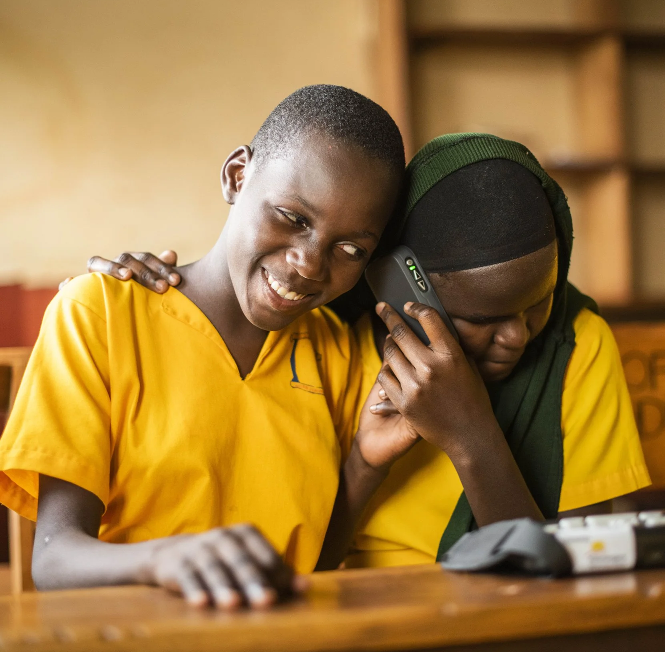 Two school-aged girls in yellow uniforms share a listening device while sitting at a desk in a classroom.