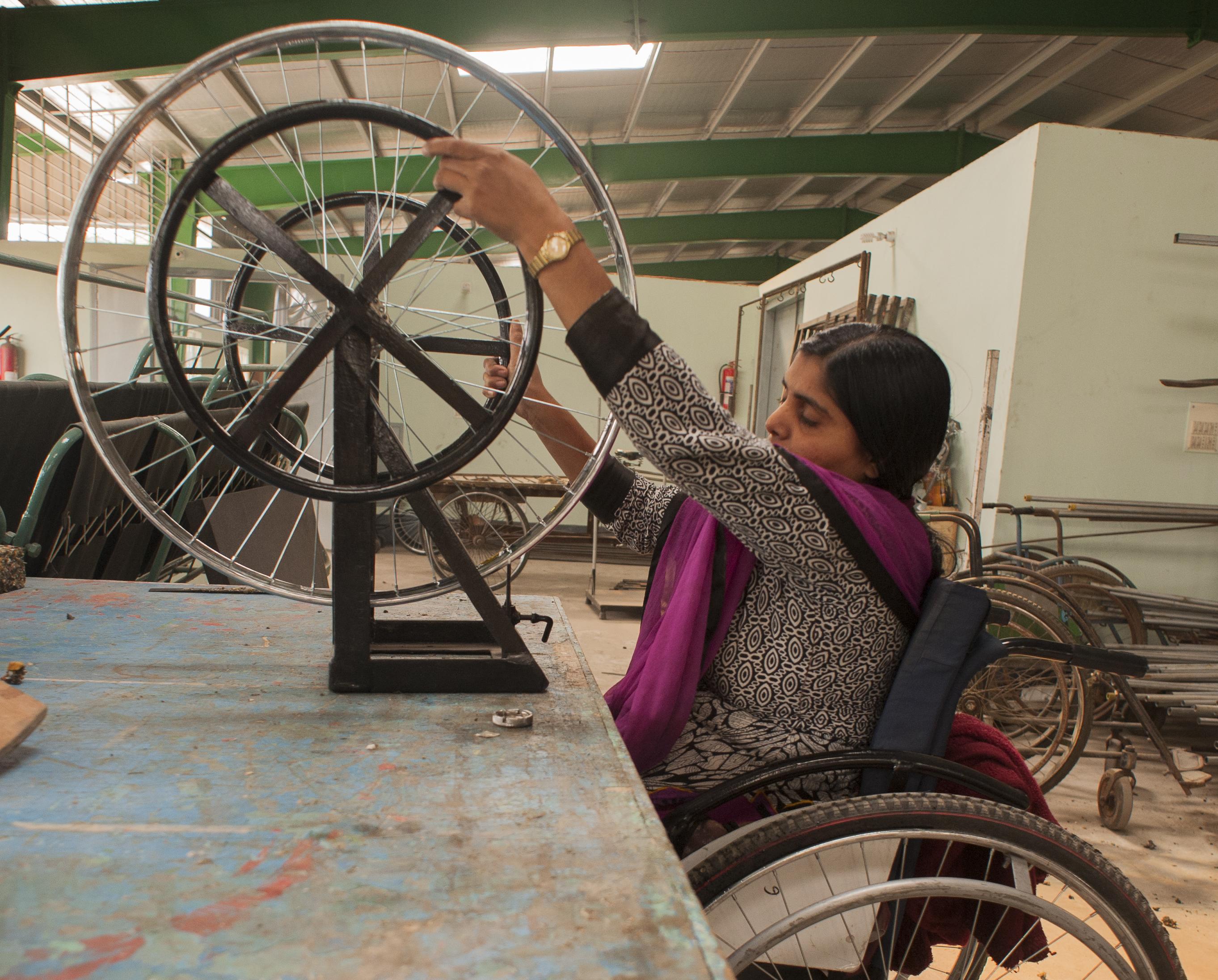 A woman using a wheelchair works on assembling a wheelchair wheel in a workshop