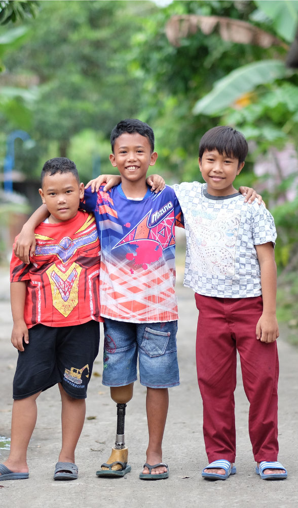 Three boys stand arm in arm on a village path, smiling; the boy in the center uses a lower-limb prosthetic