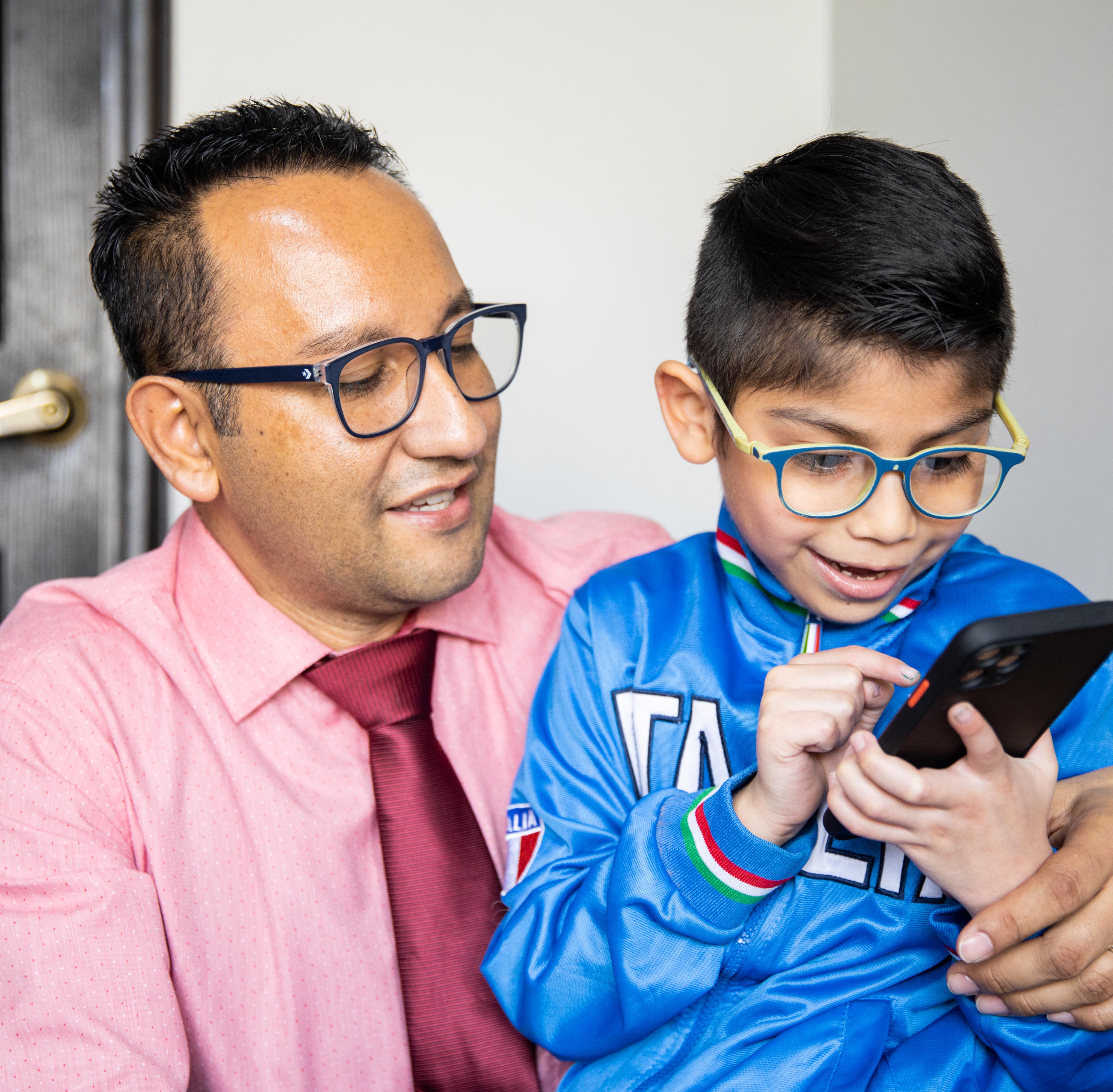 An adult sits with a child who is using a smartphone, both wearing glasses, indoors