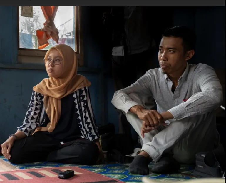 Three people sit on the floor indoors, engaged in a small group discussion inside a modest room.