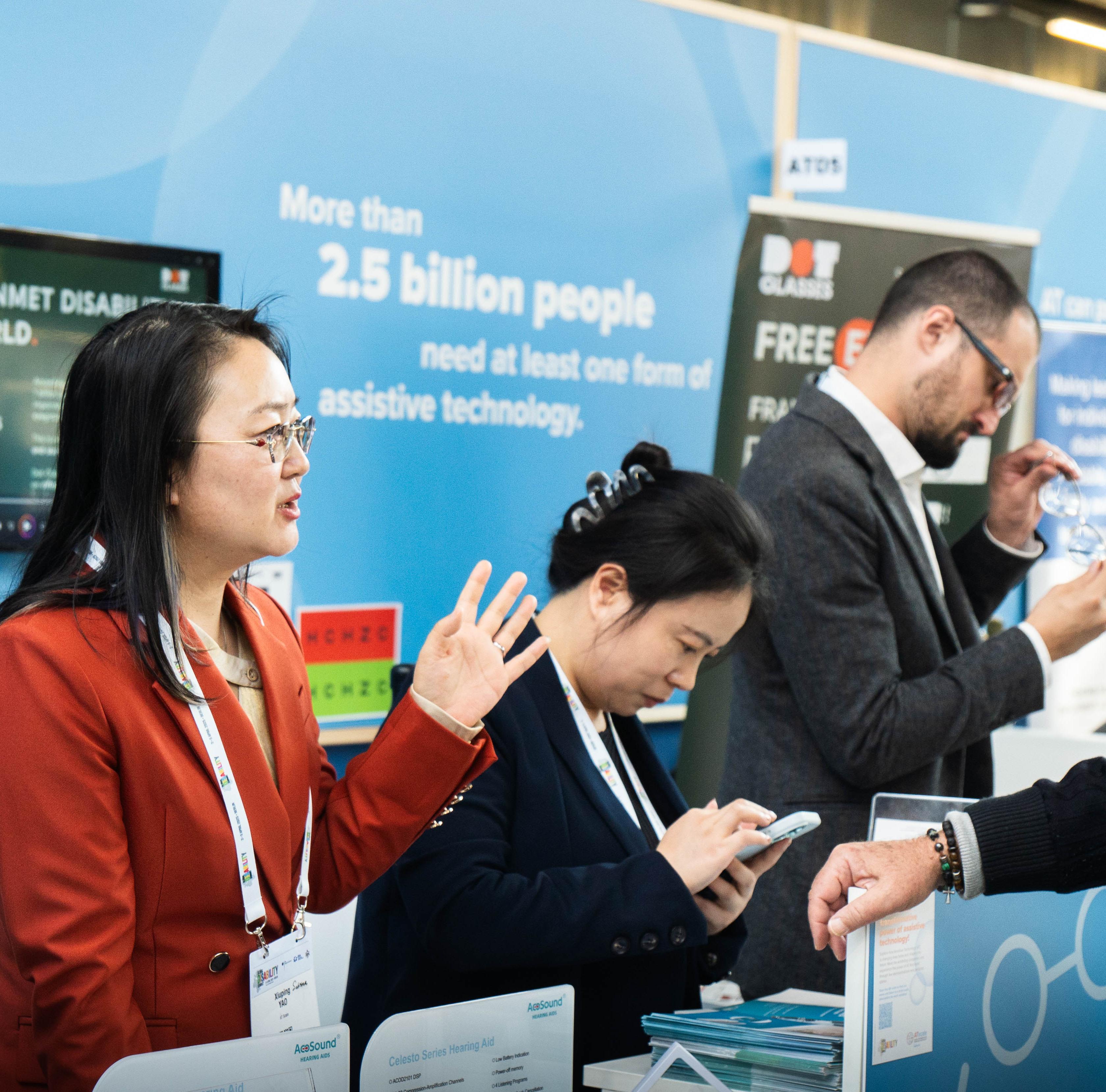 People discuss assistive technology products at an exhibition booth, with informational displays in the background