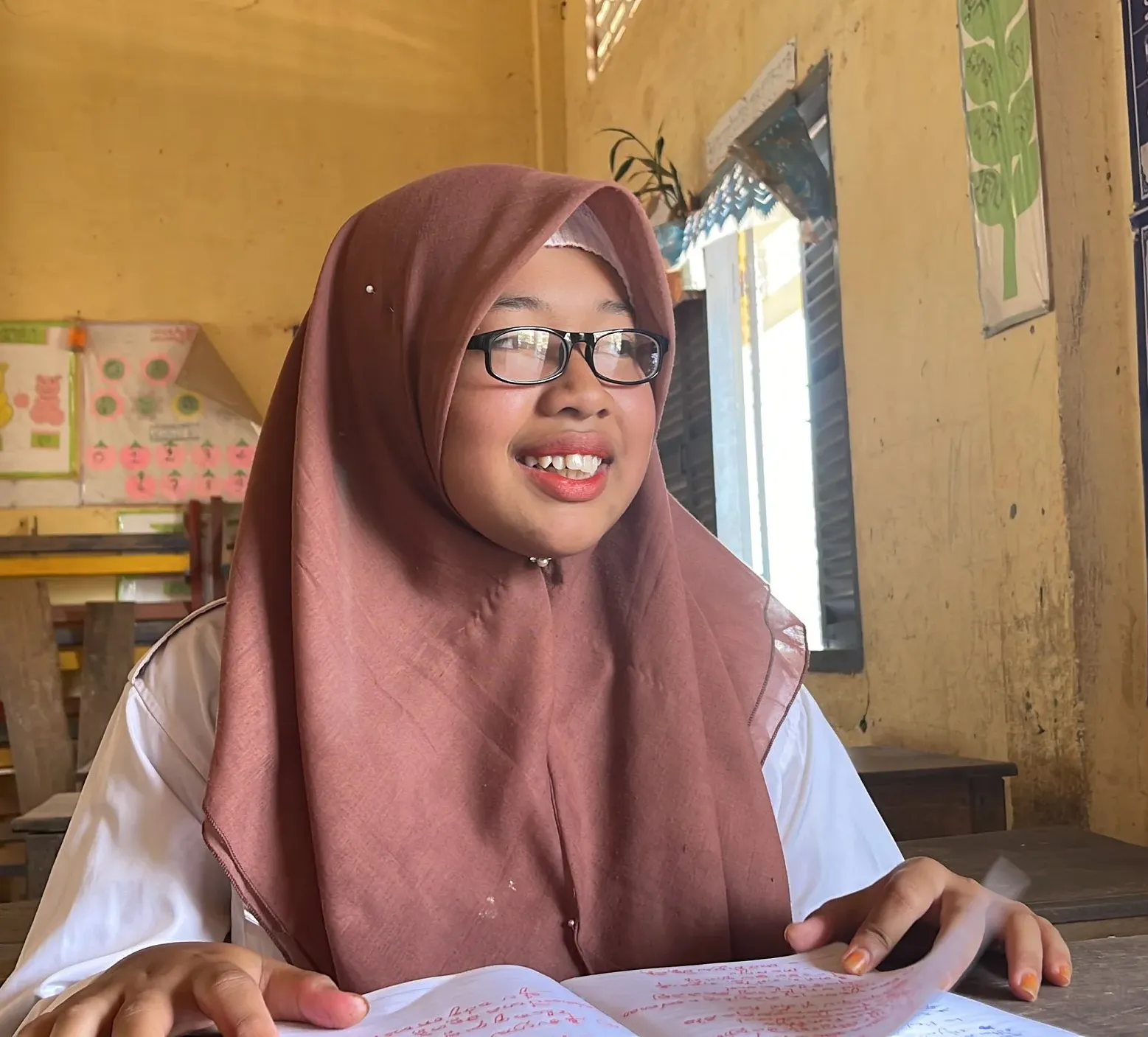 Schoolgirl wearing glasses and a headscarf sits at a classroom desk, smiling while reading from an open book