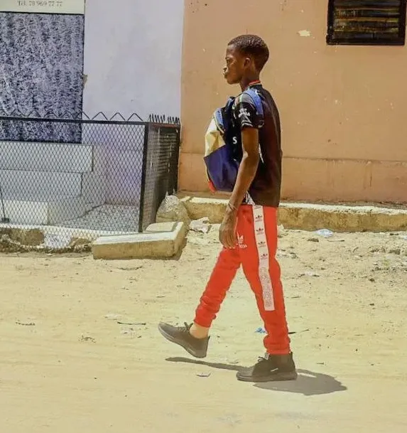Bokk Naa CiA young person walks along a dusty street in an urban neighbourhood, wearing a backpack and red trousers.