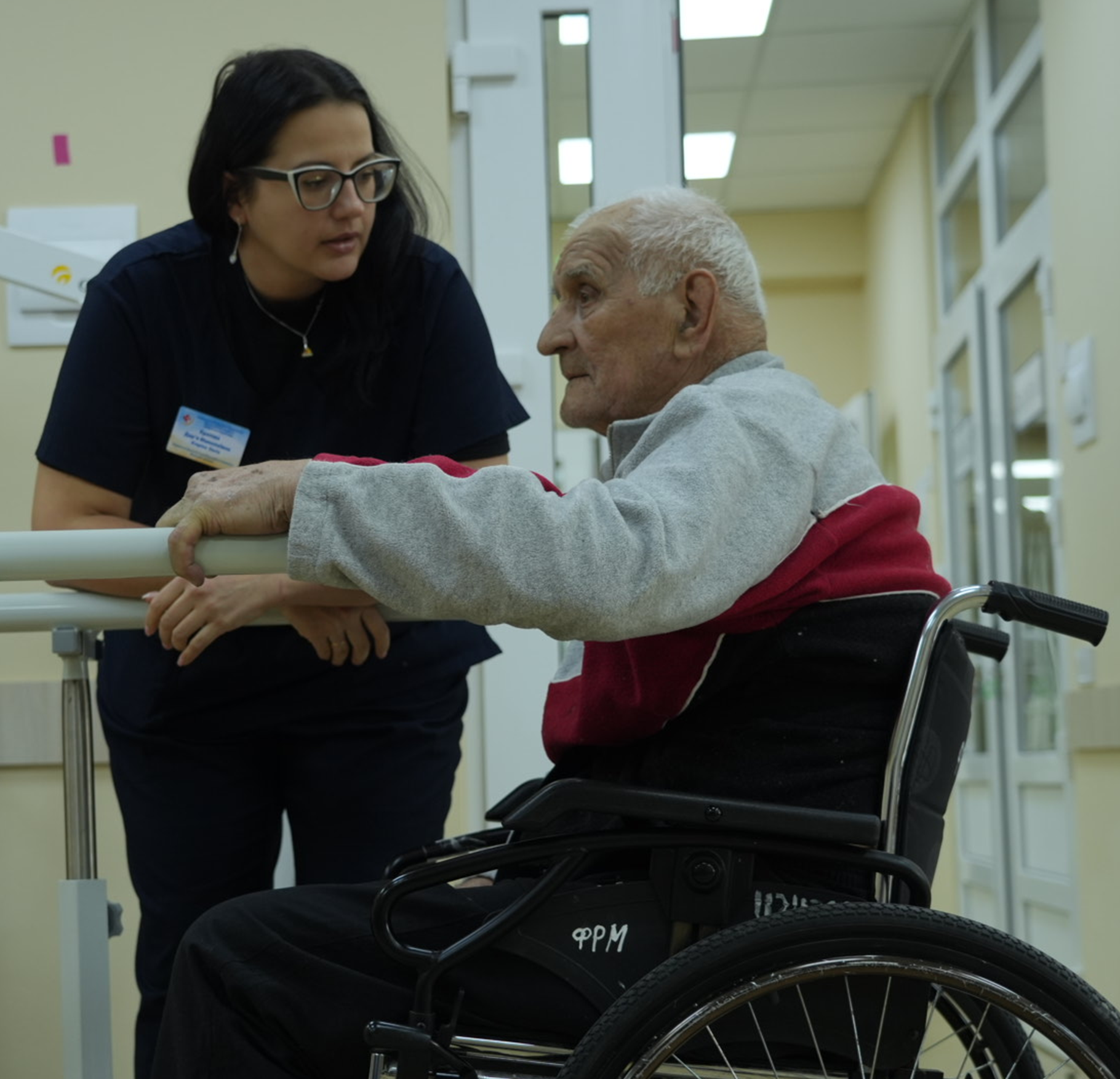 A health professional supports an older man using a wheelchair as he practices standing at parallel bars in a clinic