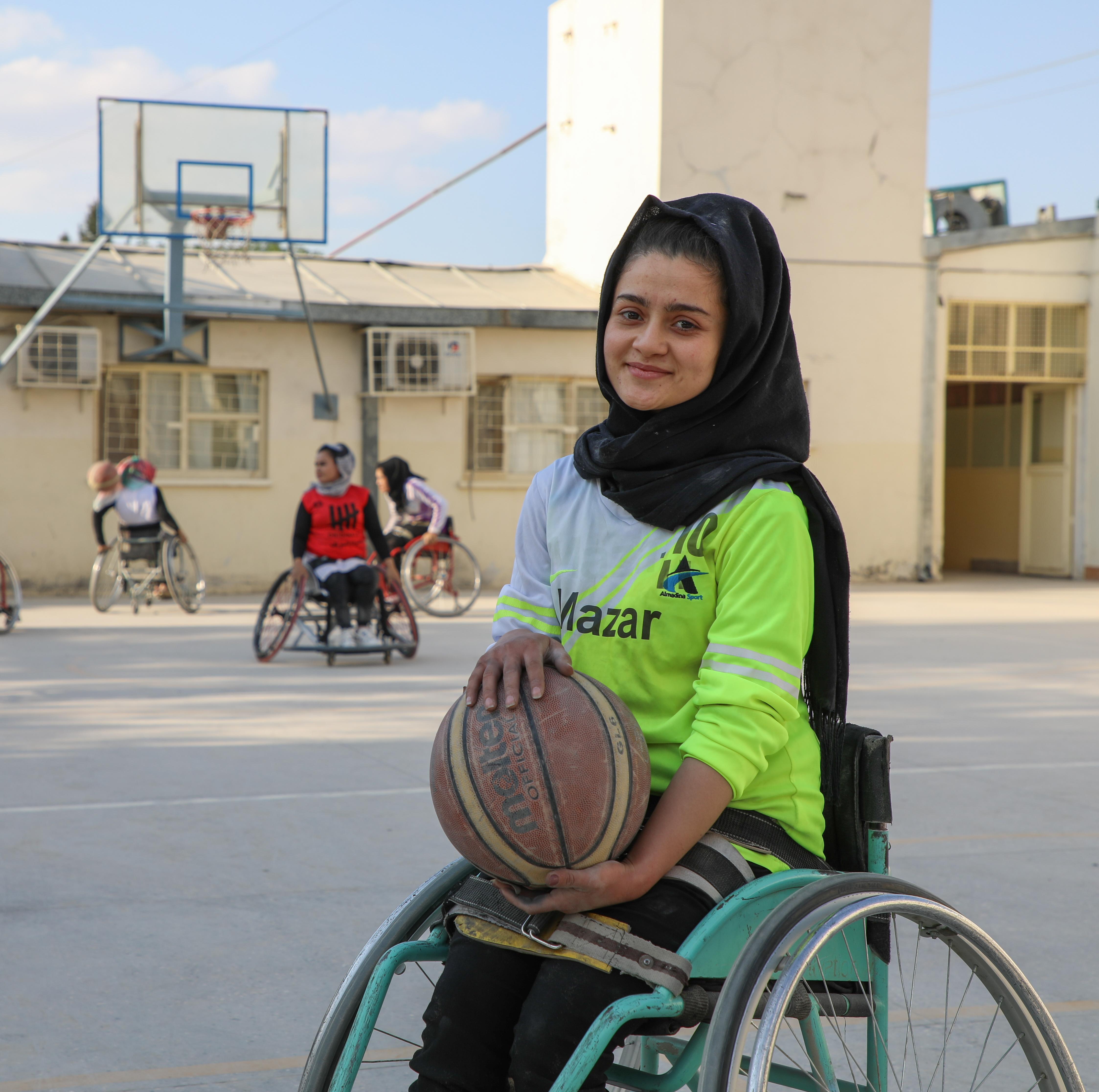 A young woman using a wheelchair holds a basketball on an outdoor court, with others playing wheelchair basketball in the background