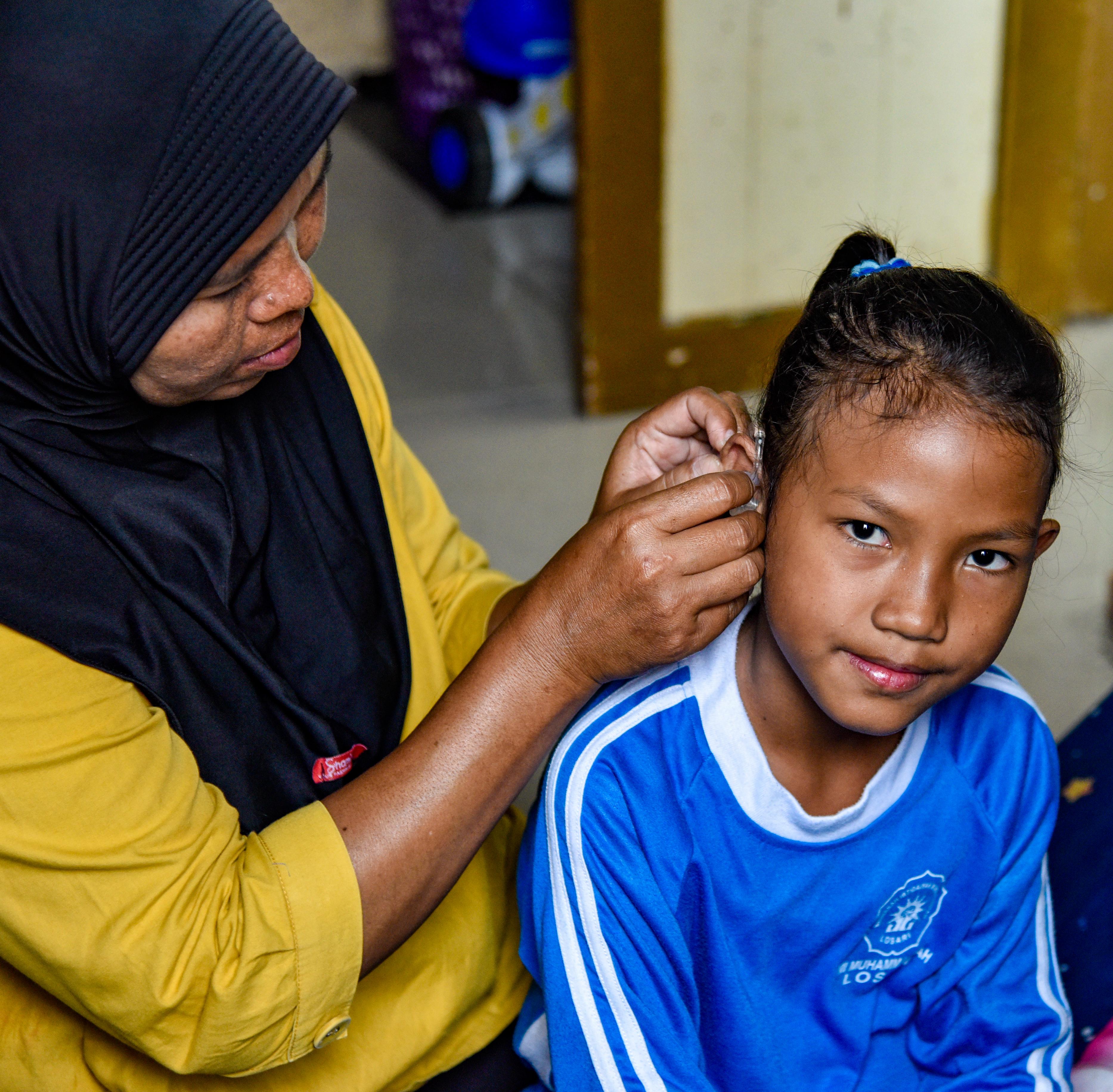 A woman helps a young girl put on a hearing aid indoors