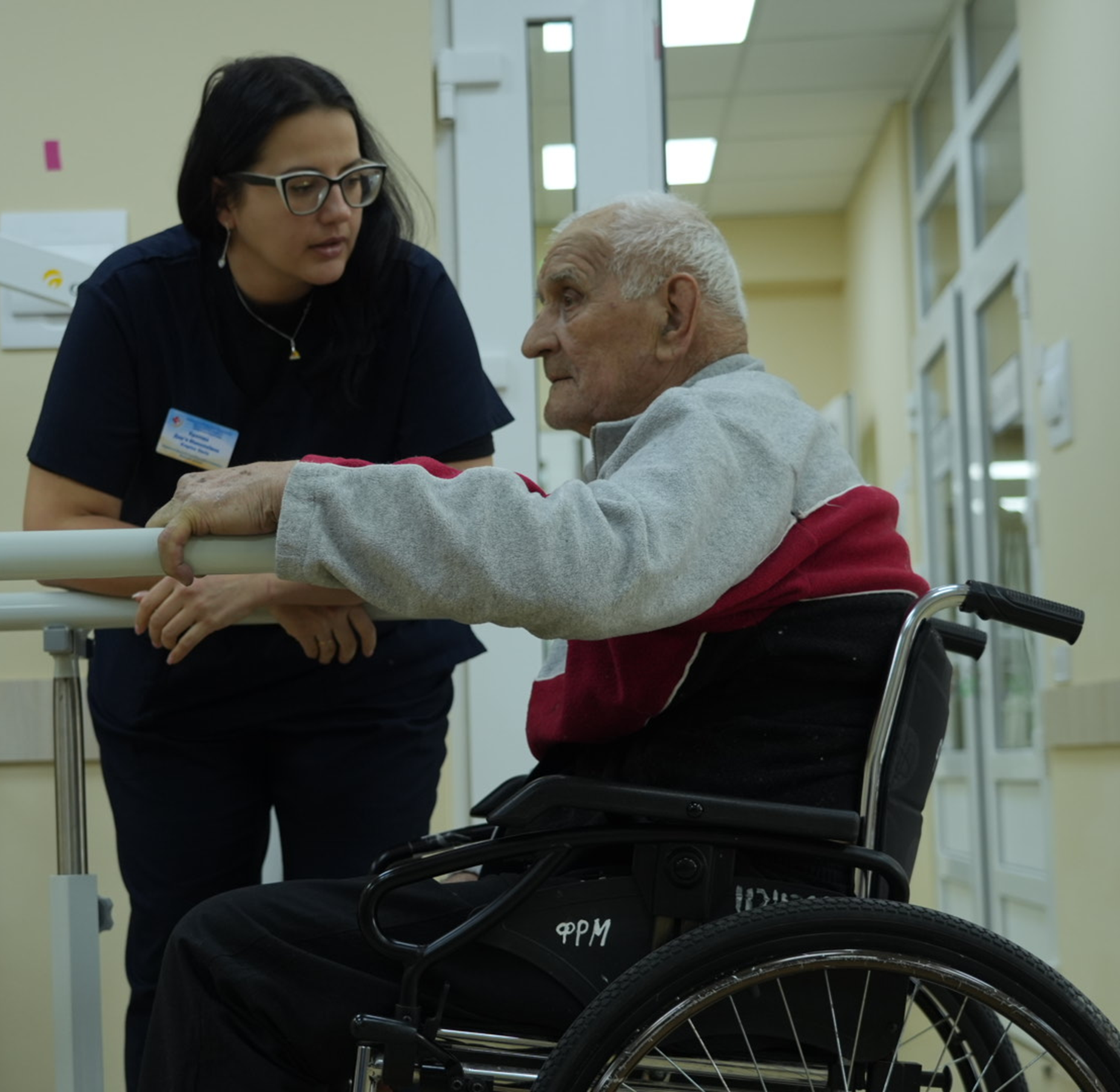 A health professional supports an older man using a wheelchair as he practices standing at parallel bars in a clinic