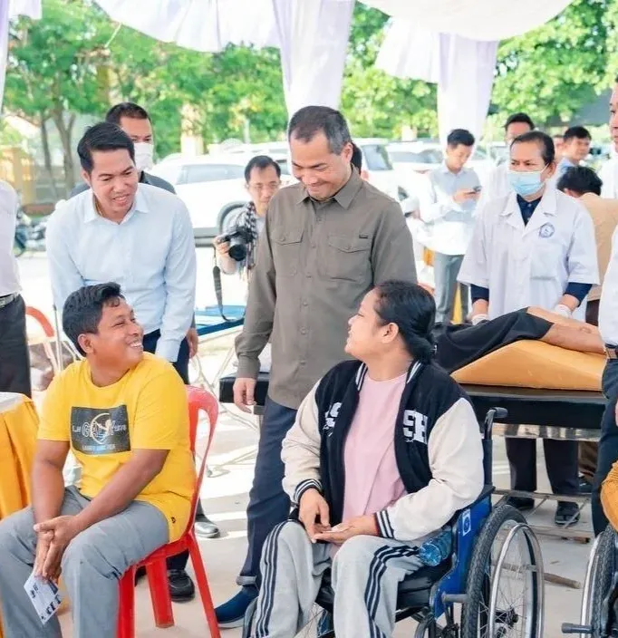 Officials and health workers speak with community members at an outdoor event; two women using wheelchairs sit among the group during a disability or rehabilitation outreach activity.