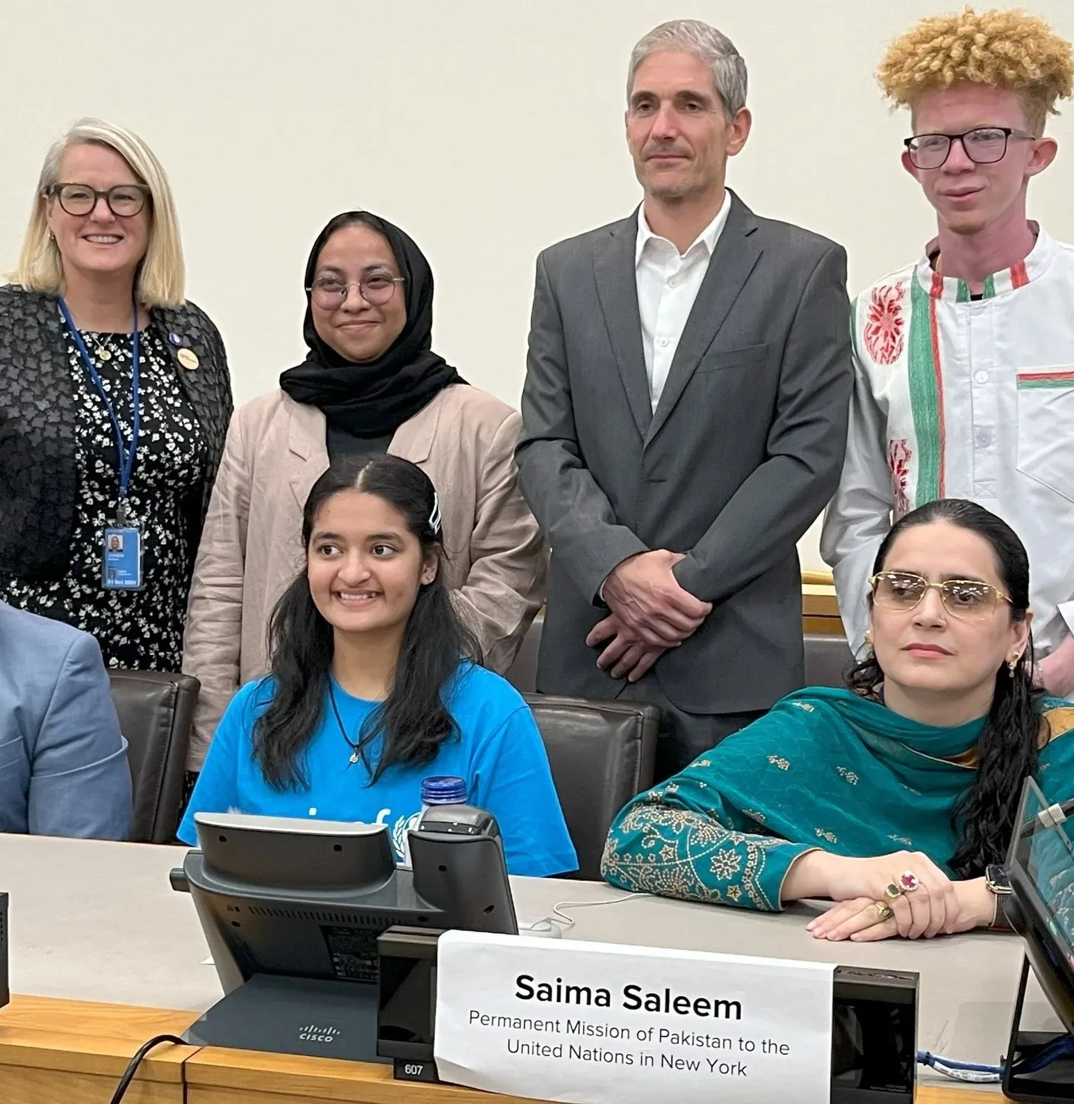 Group of delegates and advocates pose together at a conference table during a formal meeting, with nameplates visible in a United Nations setting.