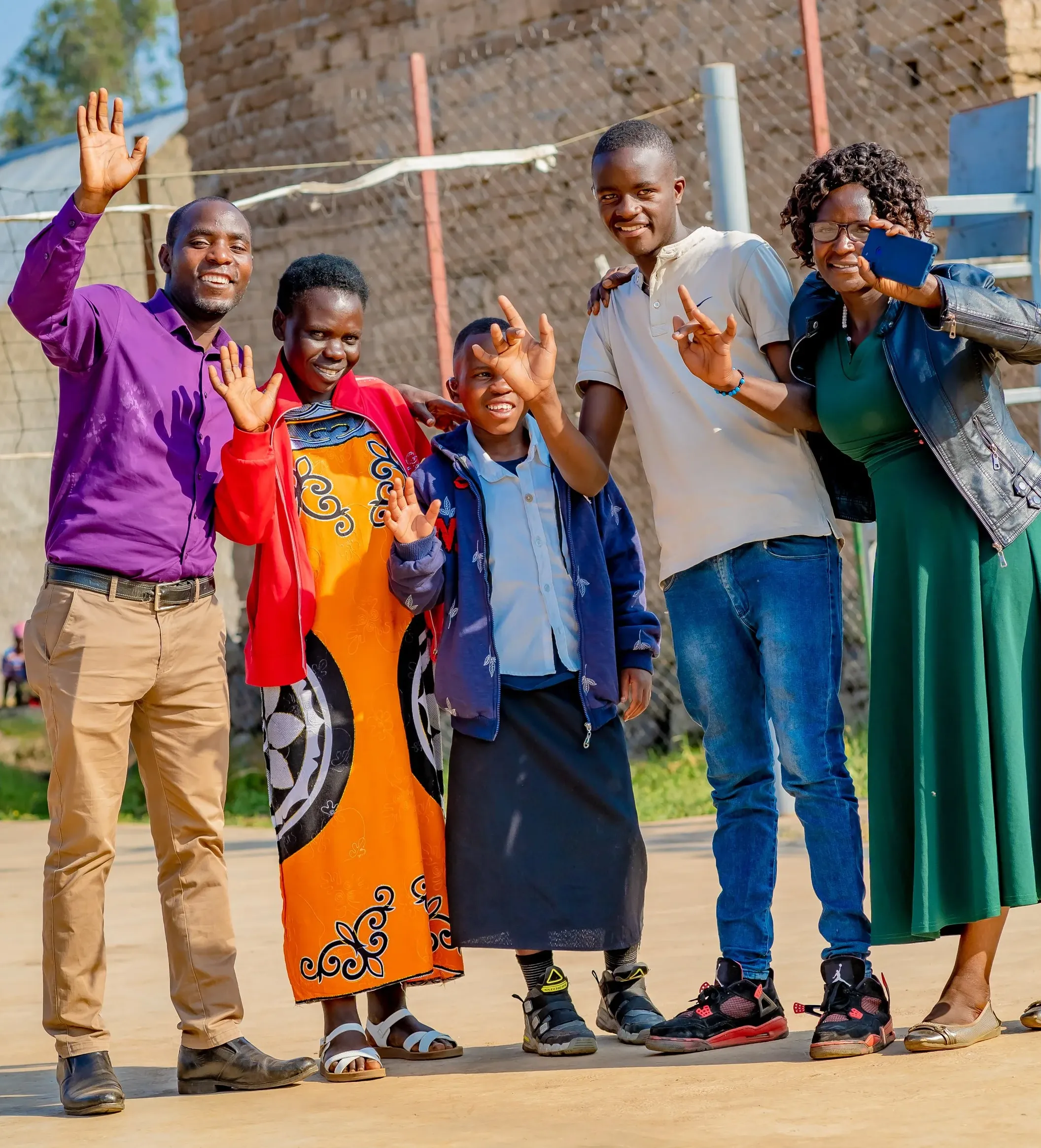 Six people of different ages stand outdoors in front of a brick building, smiling and waving at the camera in a community setting.