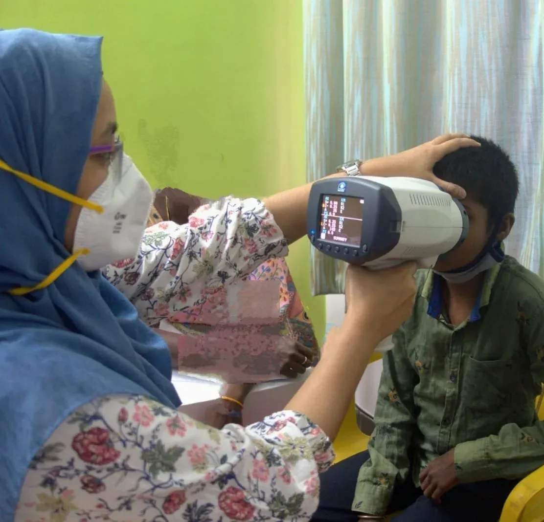 The health professional is using a hand-held autorefractor to diagnose vision impairments