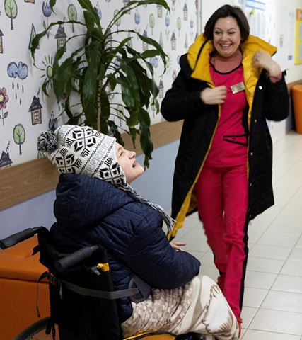 Healthcare worker smiles and speaks with a child in a wheelchair in a bright clinic hallway.