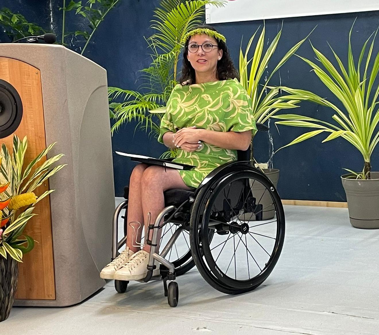 A woman in a wheelchair sits on a stage next to a podium, with potted plants in the background.