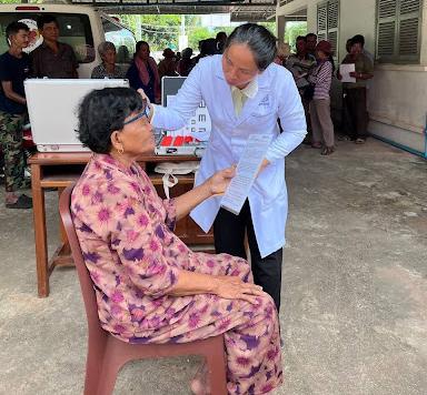 A healthcare worker in a white lab coat conducts an eye examination on a seated woman at an outdoor mobile health clinic.