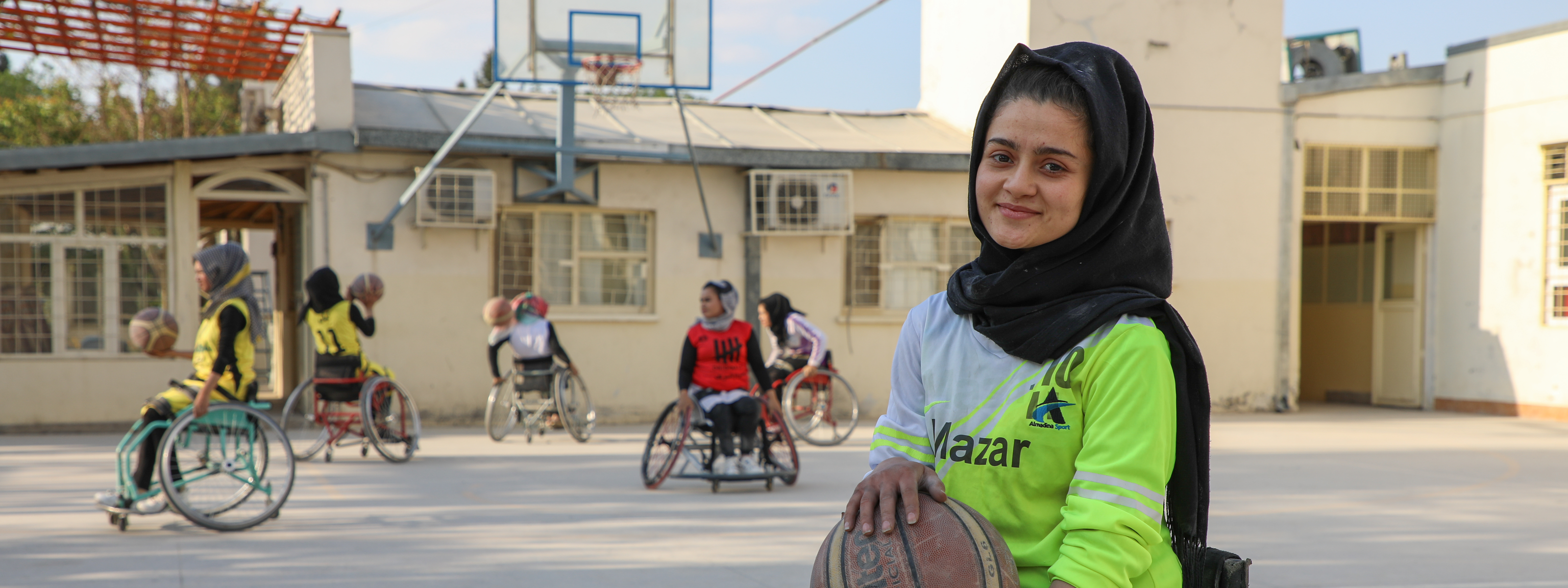 A young woman using a wheelchair holds a basketball on an outdoor court, with others playing wheelchair basketball in the background