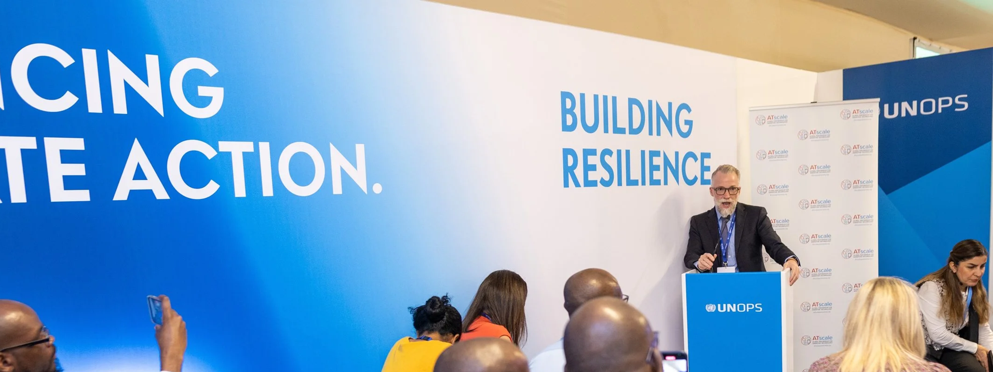 A speaker stands at a UNOPS podium addressing an audience, some using wheelchairs, during a conference session.