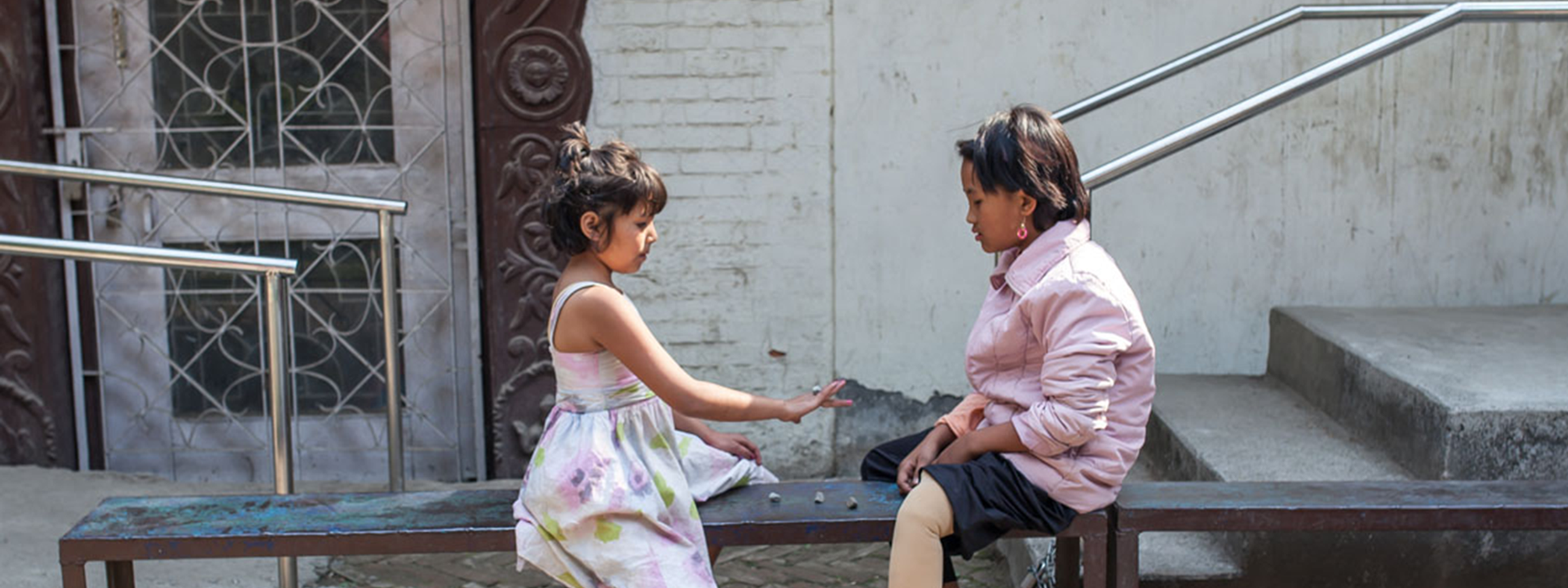 Two children sitting on a bench outdoors, both wearing prosthetic legs, talking together.