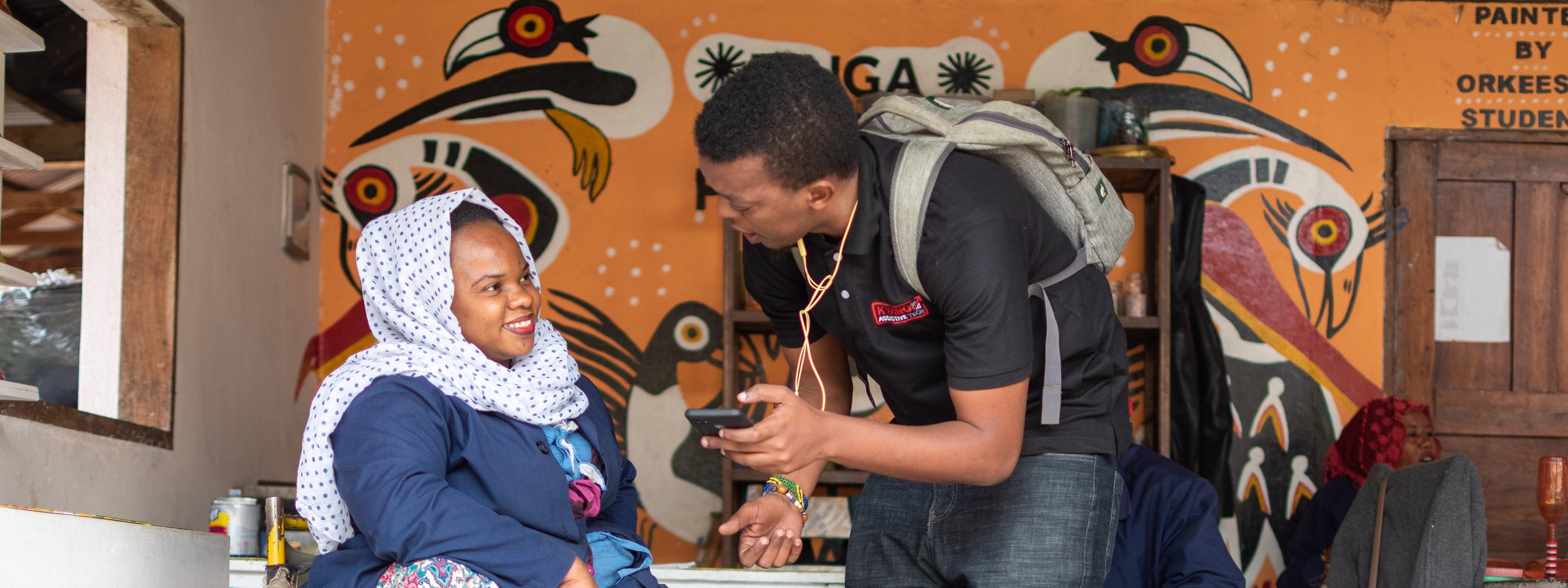 A man shows a smartphone to a woman using a wheelchair during a conversation indoors.